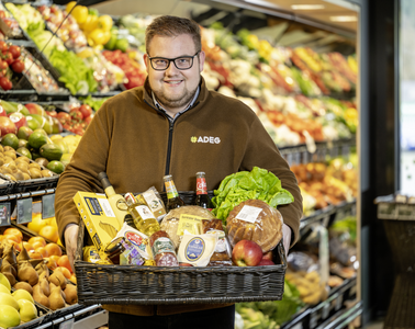 Kaufmann Benedikt mit einer Kiste voller Waren in seinem Markt