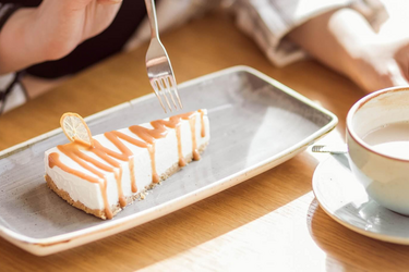 Eine Gabel sticht in ein Stück Cheesecake, daneben ein Milchkaffee