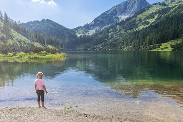Bild eines Sees in den Bergen, davor sitzt eine Frau und beobachtet ein Mädchen am Wasserrand